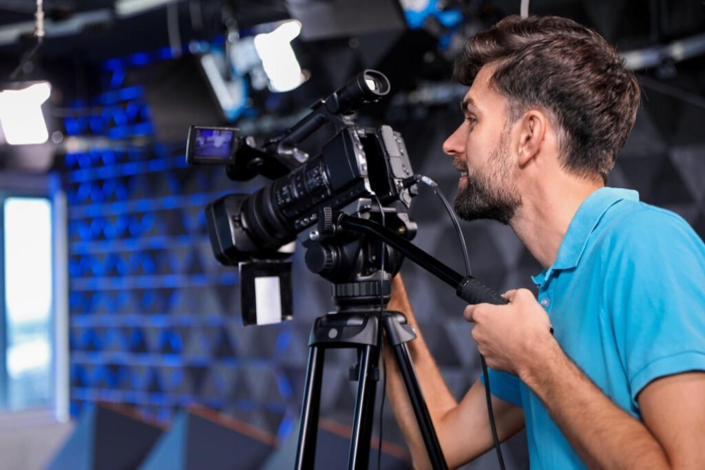 Bearded man in turquoise polo shirt operating professional video camera on tripod in broadcast studio with blue lighting and equipment in background
