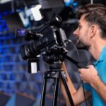 Bearded man in turquoise polo shirt operating professional video camera on tripod in broadcast studio with blue lighting and equipment in background