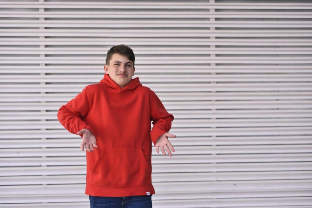 Young person wearing a bright red oversized hoodie posing confidently against a white striped metal shutter background.