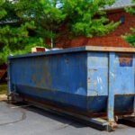 A large blue metal dumpster with rust stains and wear marks positioned on a street in front of a brick house, surrounded by green trees and lumber materials