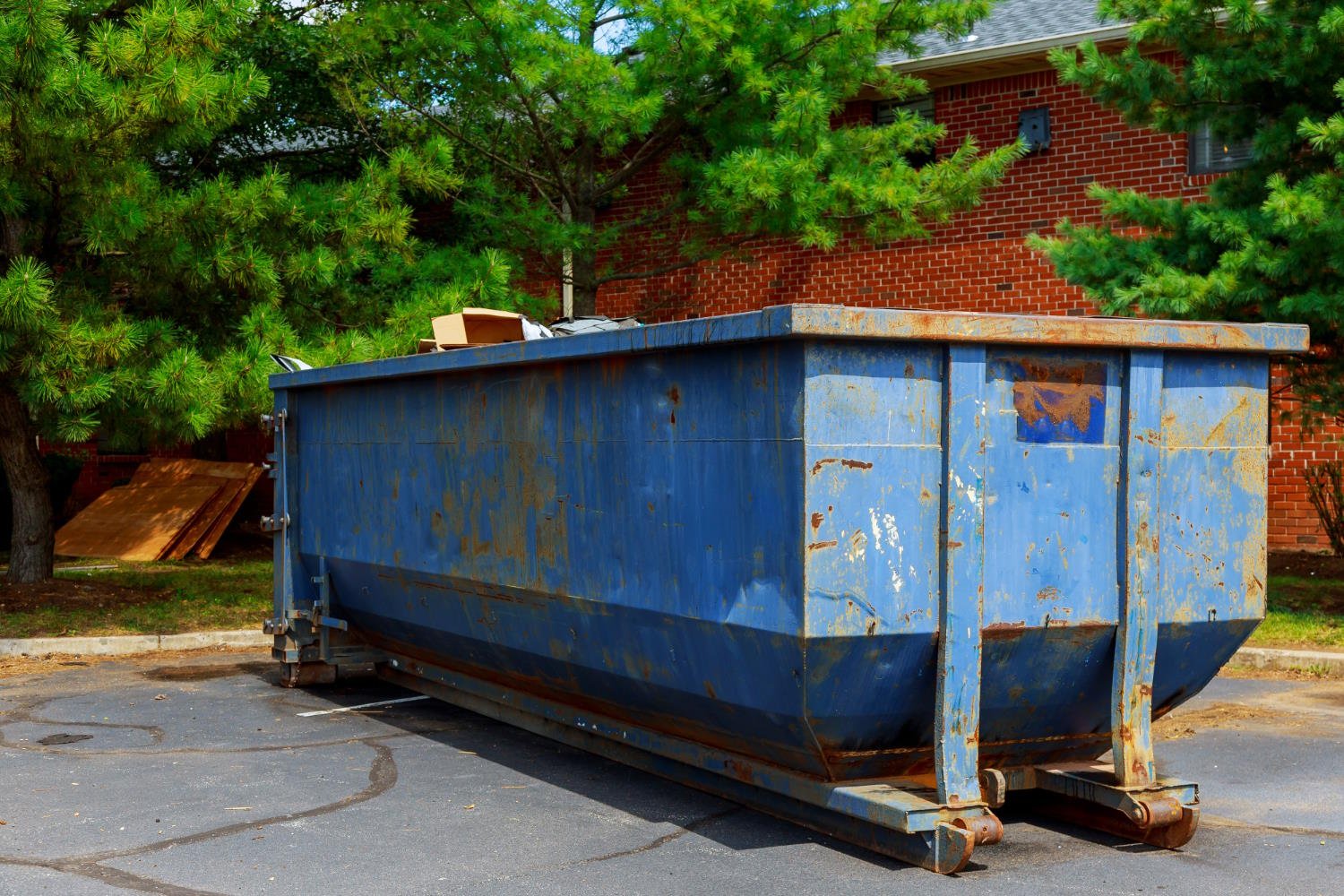 A large blue metal dumpster with rust stains and wear marks positioned on a street in front of a brick house, surrounded by green trees and lumber materials