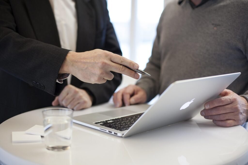Two business professionals in suits collaborating at a white desk, with one person holding a pen and pointing at documents while the other uses a laptop, with glasses and papers visible on the table.