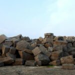 A large pile of rectangular and square-cut stone blocks in various shades of brown, tan, and gray scattered across a concrete surface against a partly cloudy blue sky. The stones appear to be quarried building materials of different sizes stacked in an irregular heap.