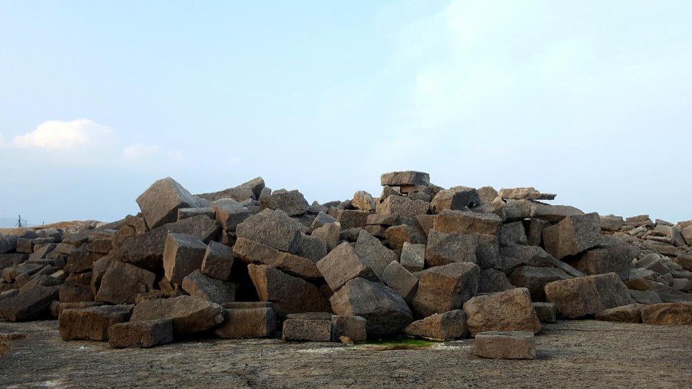 A large pile of rectangular and square-cut stone blocks in various shades of brown, tan, and gray scattered across a concrete surface against a partly cloudy blue sky. The stones appear to be quarried building materials of different sizes stacked in an irregular heap.