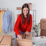 Young woman in red sweater smiling while packing cardboard shipping boxes at desk with laptop, clothing rack with colorful t-shirts visible in background of home office workspace