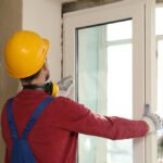 Professional window repair technician in yellow hard hat and red shirt working on white window frame during installation or maintenance