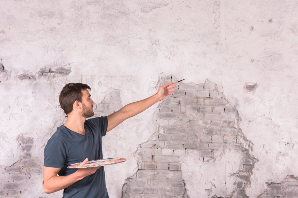 A professional contractor in a dark blue shirt holding a clipboard and pen, pointing to and examining severely damaged interior walls with peeling paint, exposed brick, and water damage stains, demonstrating the thorough assessment process required for flood-damaged building restoration.